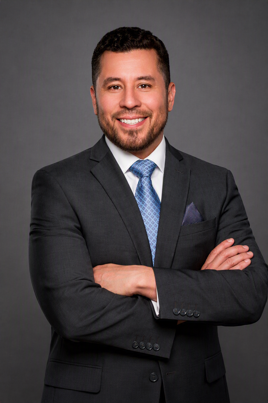 Louis Oliveira smiling in a charcoal suit with a blue patterned tie against a gray studio background